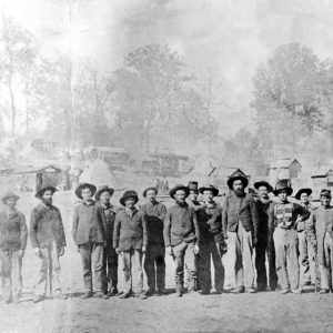 Soldiers posing in line wearing hats with buildings cabins tents on hillside in background