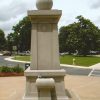 Stone monument with stone sphere on top and water fountain on its base with driveway and trees behind it