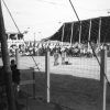 men and horses in outdoor arena with covered stands behind fencing as seen from opposite stands