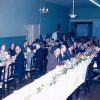 Group of older white men and women with glasses in formal clothing eating at long tables in dining hall