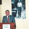 White man in suit speaking "W.R." logo lectern by portrait of white man with automobile