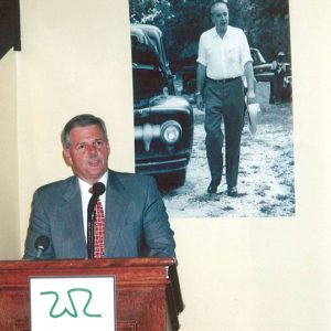 White man in suit speaking "W.R." logo lectern by portrait of white man with automobile