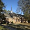 Side view of single-story brick church building with covered porch and steeple with tall tree next to it