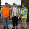 Two white men and woman in reflective vest standing by river with concrete bridge and city buildings in the background