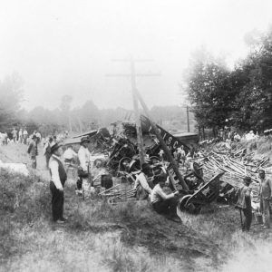 Mixed spectators gathered around wrecked train cars and their spilled cargo with trees in the background