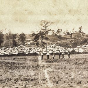 Group of men standing in field with rows of tents behind them