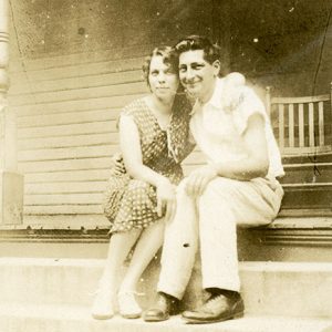 White man and woman sitting on porch with chair behind them