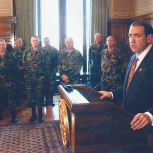 White man at lectern speaking in room alongside several people in camouflage