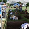 many white people stand around and in square shaped holes in the ground with white marquee tents providing some shade