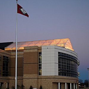 multistory brick building with glass ceiling with lights showing through and two flagpoles outside with American and Arkansas flags