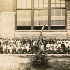 large group of people posing with large telescope in front of multistory stone and brick building