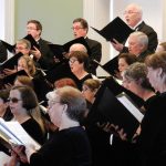 Group of white men and women singing in choir with sheet music books in hand