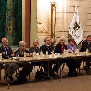 Group of white men and women sitting at tables with water bottles in large room with flag and brick wall behind them