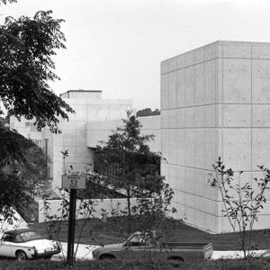 Row of cars and truck parked outside multilevel concrete and glass building