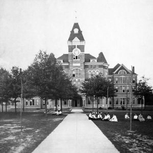 Students seated in school uniforms on quad outside large multistory brick building with clock tower