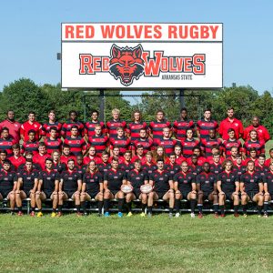 Group of young men and coaches in red and black  uniforms in front of "Red Wolves Rugby" sign