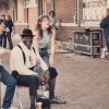 African-American man in white shirt suspenders and hat sitting and laughing with white women posing with him outside brick building