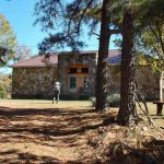 Man with cowboy hat standing outside stone building with bench and trees in the foreground