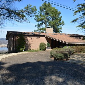 Single-story house with angled flat roof and covered entrance with chimney and river in the background