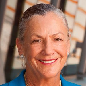 Older white woman smiling in blue suit with earrings and pendant necklace