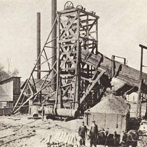 White men standing before full rail car parked by large mine shaft