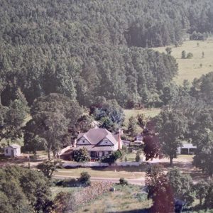 Multistory house and outbuildings on dirt road surrounded by tree covered countryside view from above