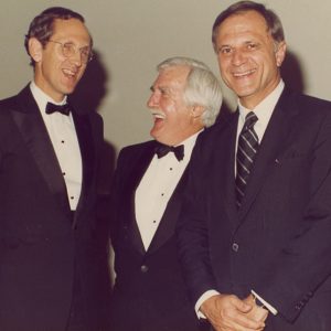 White man in suit and tie smiling while white men in suit with glasses and older white man in suit laugh behind him