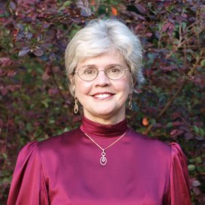 smiling white woman with gray hair wearing glasses earrings in red dress with necklace