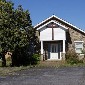 Single-story church building with cross on covered porch and stone walls with steeple in the background