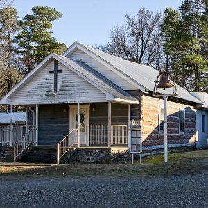 Single-story church building with covered porch log walls and bell in the front yard