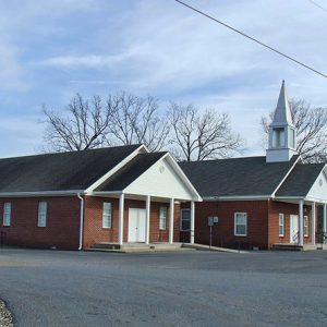 Single-story brick church buildings one with steeple and sign on parking lot