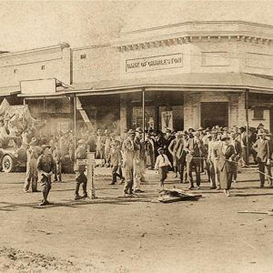 Large crowd of people standing on street with storefronts in the background