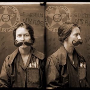 White woman with curly mustache and cigars in her shirt pocket with sheriff's office logo behind her