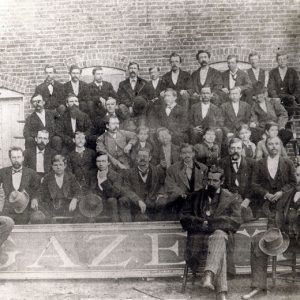 Group of white men in suits posing outside a brick building with large sign "Gazette" at their feet