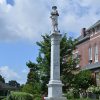 statue of soldier with hat and gun on top of stone monument with pedestal inside iron fence next to multistory brick building with gazebo