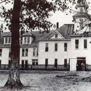 wide white building with gable roofs and bell tower with trees in foreground