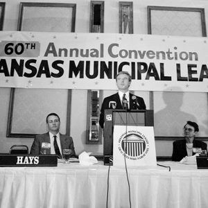 White men in suits seated at long table with white man in suit speaking at lectern with banner hanging behind them