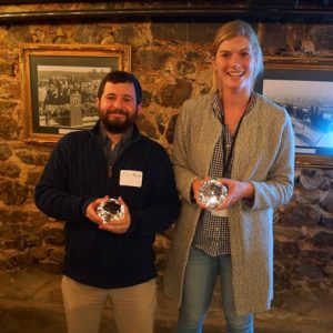 White man with beard smiling with award standing next to white woman in long coat smiling with award