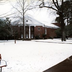 Snow covered quad and bench with crossing sidewalks leading to large multistory neoclassical brick building with white columns