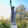 Statue of woman in robes and crown with torch in her right hand and tablet in her left on stone pedestal with plaque on it between two bushes