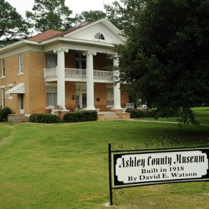 two story yellow brick house with white columns and balcony on green lawn with trees and sign reading "Ashley County Museum Built in 1918 by David E Watson"