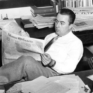 White man sitting at his desk reading a newspaper with typewriter behind him and stacks of newspapers around