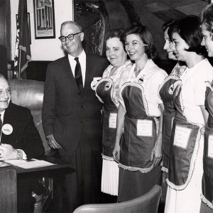 Two white men in suit and tie with five white women wearing aprons in office