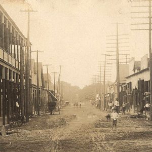 Multistory buildings and telephone poles on either side of dirt road with white child standing in it