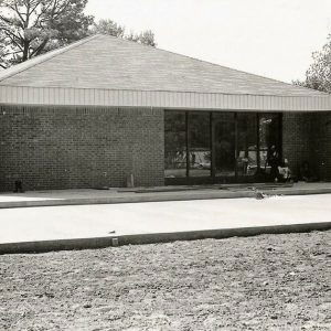 Man on ladder outside single-story brick building with pyramid roof and tall glass windows around glass door