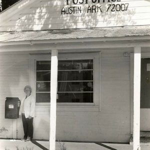 Old white woman standing by mailbox outside single-story post office building with covered porch