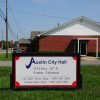 Street with "Austin City Hall" sign and brick building with gas station in the background