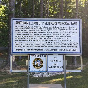 "American Legion B-17 Veterans Memorial Park" sign with text and smaller signs on grass with trees behind them