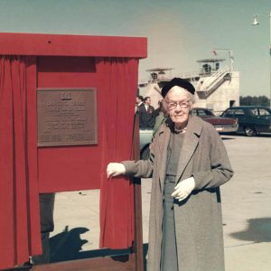 Old white woman standing next to dedication plaque with parked cars and crowd behind her