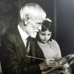 Old white man in suit and tie at easel with young white girl next to him